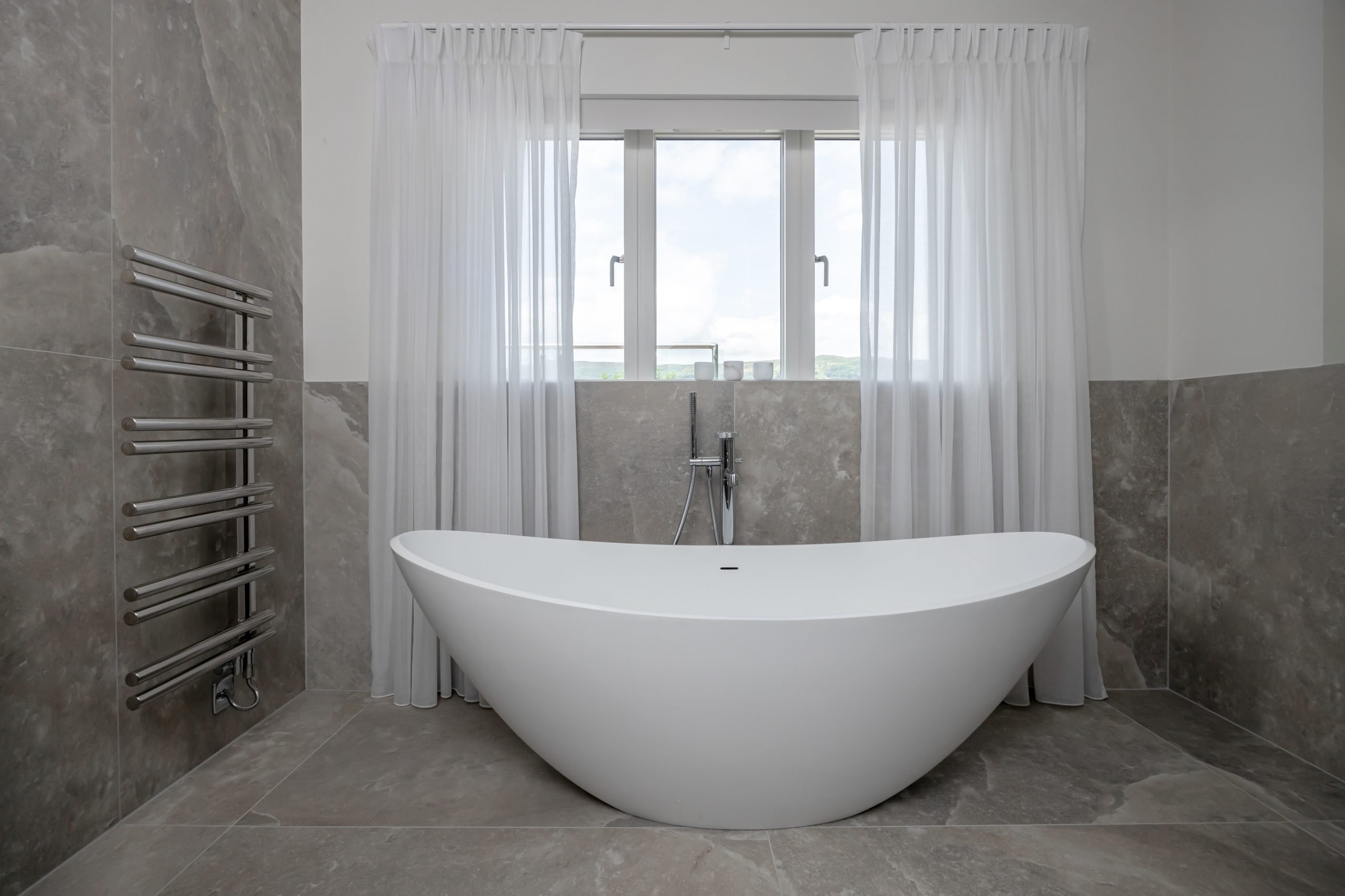 Sculptural freestanding white bathtub framed by floor‑to‑ceiling grey stone tiles and sheer curtains in modern Welsh bathroom